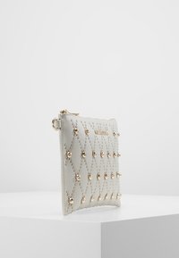 Pochette en cuir blanc avec un design matelassé orné de clous dorés, une texture lisse et une fermeture éclair. Dimensions non spécifiées.