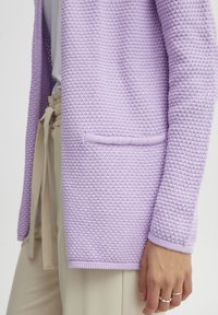 Lila stickad kofta med ett texturerat mönster, långa ärmar och en framficka. Kompletteras av ljusbeiga byxor.