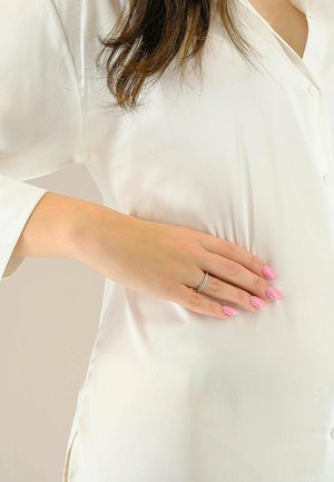 Blusa bianca e setosa con colletto standard e maniche lunghe. Una mano con un anello d'argento e unghie rosa poggia sul tessuto.