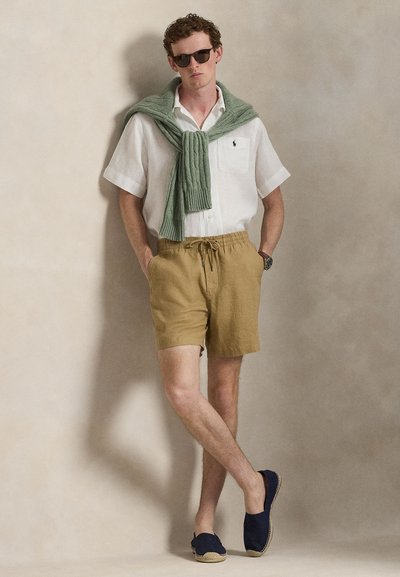 Ελαφρύ λευκό πουκάμισο από λινό με κοντά μανίκια, καφέ σορτς με κορδόνι, πράσινο πλεκτό πουλόβερ δεμένο στον λαιμό, μπλε σκούρα παπούτσια τύπου slip-on, σκούρα γυαλιά ηλίου.