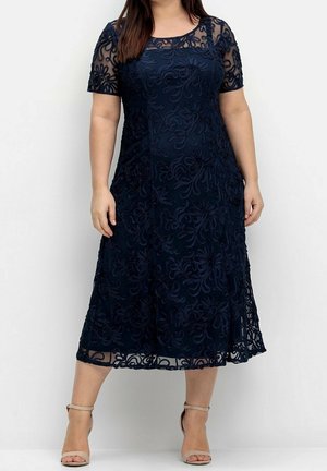 Vrouw die een marineblauwe, knielange jurk draagt met een bloemenkantlaag en korte mouwen, gecombineerd met beige enkellaarzen met riempje.