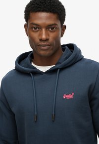 Marineblauwe hoodie met trekkoorden en een roze geborduurd logo. Gemaakt van zachte stof, met een kangoeroezak en geribde manchetten.