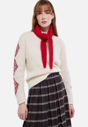 Femme portant un pull crème avec des motifs de losanges rouges sur les manches, une écharpe rouge tricotée, et une jupe plissée à carreaux marron foncé.