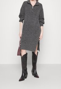 Robe pull en tricot gris avec des poignets côtelés, un col zippé mi-haut et des fentes latérales, assortie à des bottes de cowboy noires brillantes et un sac bordeaux.