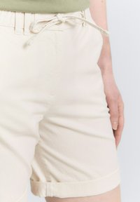 Pantalones cortos de algodón beige con un acabado texturizado, que cuentan con una cintura ajustable con cordón y puños enrollados. Los bolsillos laterales visibles añaden practicidad.