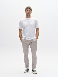 Camiseta blanca de manga corta con logo negro, pantalones ajustados beige claro y zapatillas a rayas en blanco y negro. Atuendo simple y casual.