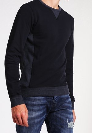 Mann trägt geschichtete schwarze und dunkelgraue Langarmshirts und ausgewaschene blaue Jeans mit einem kleinen Riss am rechten Oberschenkel.