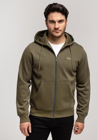 Oliwkowa bluza z kapturem na zamek, wykonana z miękkiego materiału, z kieszenią z przodu, kapturem ze sznurkiem i subtelnym logo na piersi.