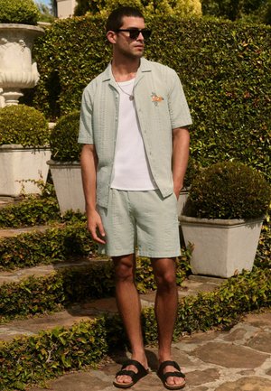 Man wearing light green short-sleeve shirt and matching shorts with orange embroidery, white t-shirt, sunglasses, and black sandals outdoors.