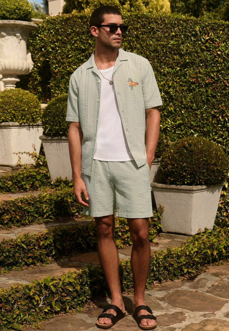 Homme portant une chemise à manches courtes vert clair et un short assorti avec une broderie orange, un t-shirt blanc, des lunettes de soleil et des sandales noires en plein air.