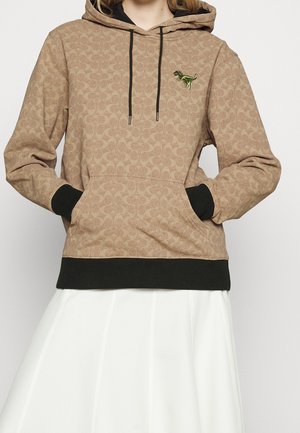 Personne portant un sweat à capuche beige avec des poignets et un ourlet noirs, un patch dinosaure sur la poitrine, les mains dans la poche avant, et une jupe blanche.