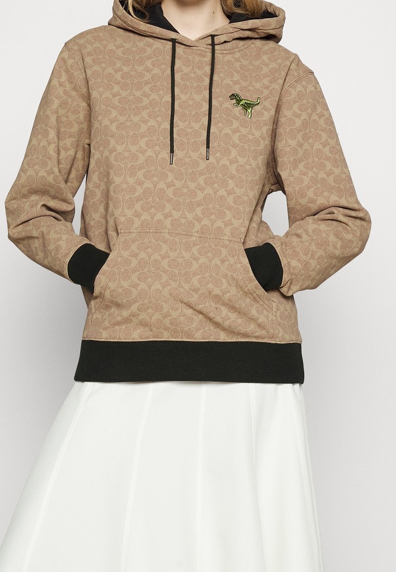Personne portant un sweat à capuche beige avec des poignets et un ourlet noirs, un patch dinosaure sur la poitrine, les mains dans la poche avant, et une jupe blanche.