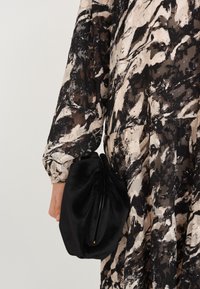 Sac à main en velours noir avec un design plissé tenu à la main, associé à une robe à manches longues à motif abstrait dans des nuances de noir et de beige.