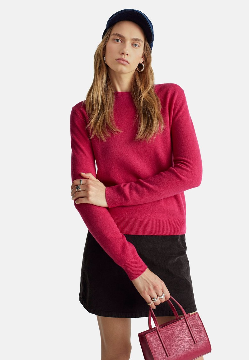 Maglione aderente a maniche lunghe di un vivace rosa, realizzato in materiale morbido. Abbinato a una gonna nera e a una piccola borsa rossa con texture.