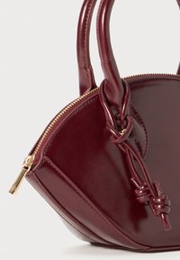Bordeauxfarbene Lederhandtasche mit glänzender Oberfläche, runder Form, doppelten Griffen und einem goldenen Reißverschluss. Besitzt eine geknotete Akzentuierung an der Seite.
