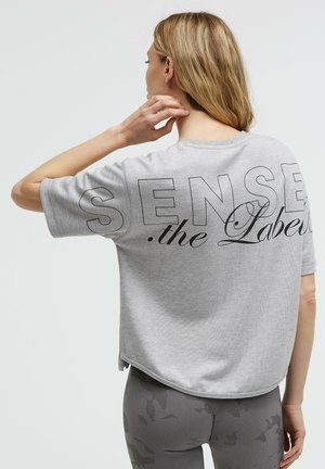 Person mit schulterlangem blondem Haar, die ein hellgraues, kurz geschnittenes T-Shirt mit der Aufschrift "SENSE the Label" und graue, gemusterte Leggings trägt, von hinten gesehen.
