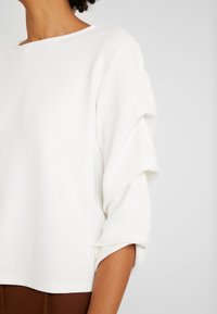 Kvinna klädd i en vit, texturerad långärmad topp med rynkade ärmar, tillsammans med bruna byxor mot en neutral bakgrund.