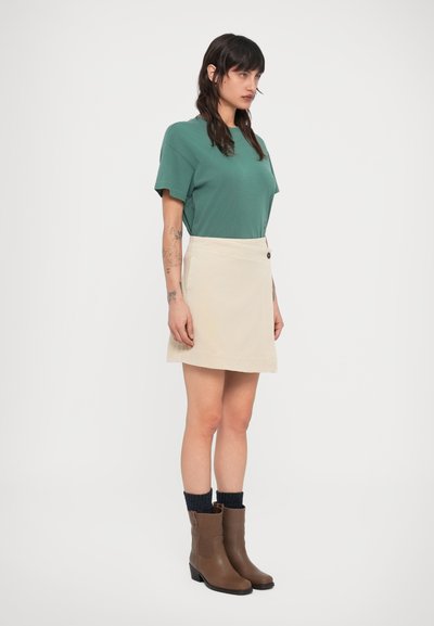 Μπεζ φούστα τύπου wrap με κουμπί, συνδυασμένη με πράσινο βαμβακερό μπλουζάκι. Καφέ μποτάκια αστραγάλου με χαμηλό τακούνι και μαύρες πλεκτές κάλτσες.