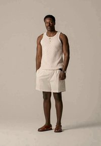 Man wearing sleeveless beige knit top, light beige shorts, brown sandals, watch, and layered necklaces standing against plain background.