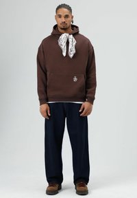 Bruine capuchonsweatshirt met voorzak, witte bandana als neksieraad, losse donkere spijkerbroek en bruine veterlaarzen.