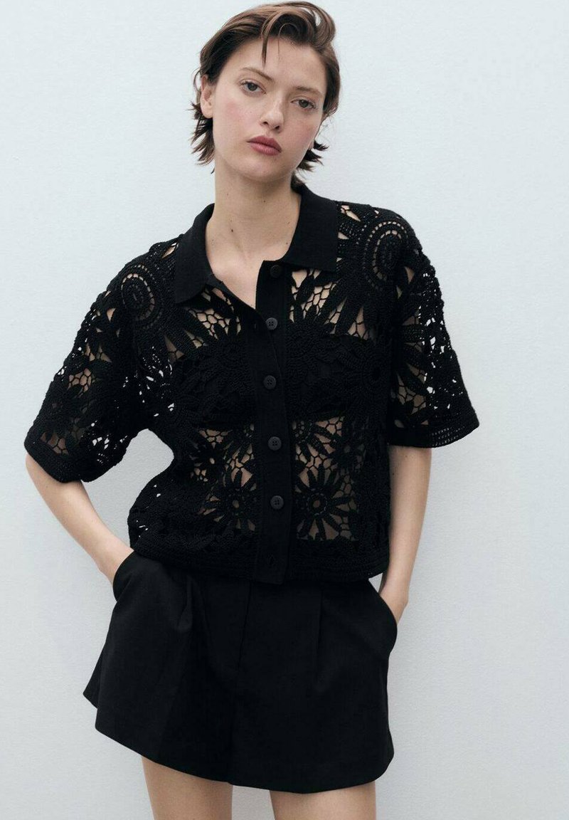Black lace blouse with short sleeves and button-down front, featuring floral patterns and a contrasting solid black collar. Paired with black shorts.