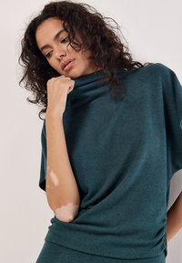 Turkos oversized tröja med hög polokrage. Mjuk, slät textur med en lös passform. Noterbara detaljer inkluderar rynkad tyg vid midjan.