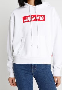 Felpa bianca con cappuccio, dotata di tasca a canguro frontale e cordini. Presenta un logo rosso di Levi's sul petto. Tessuto dalla trama liscia.