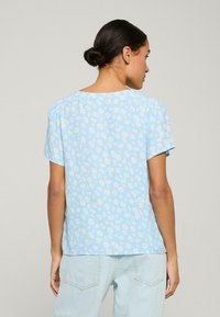 Světle modré tričko s krátkým rukávem, zdobené potiskem sedmikrásky, s kulatým výstřihem a mírně nabíranými rukávy. Látek vypadá měkce a lehce.
