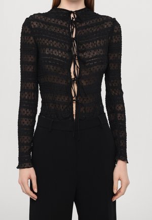 Femme portant un haut noir transparent à manches longues avec des rayures horizontales et des fermetures à nouer à l'avant, associée à un pantalon noir taille haute.