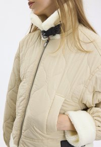 Veste matelassée beige avec un col haut, dotée d'une sangle à boucle noire, d'une doublure en polaire crème douce et de poches latérales.