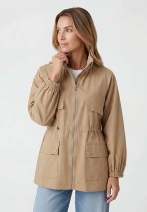Vrouw die een beige jas met rits, elastische boorden en grote voorzakken draagt, kijkt opzij tegen een effen achtergrond.