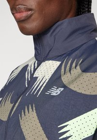 Granatowa wiatrówka z wysokim kołnierzem, abstrakcyjnymi wzorami w kolorze kremowym i oliwkowym oraz małym białym logo New Balance na piersi.