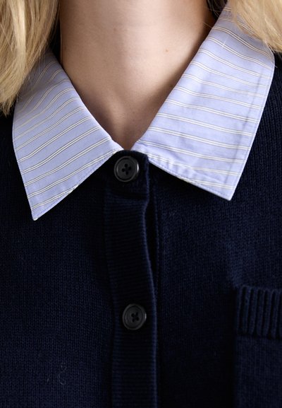 Cardigan a maglia blu navy con una camicia colletto a strisce blu chiaro sotto, dotata di un bottone nero al collo e piccolo dettaglio della tasca.