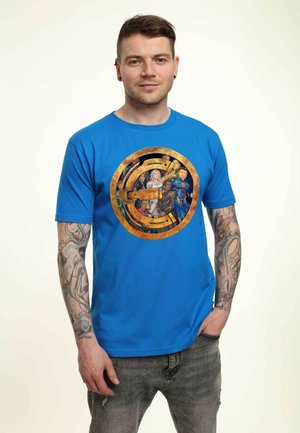 Niebieska bawełniana koszulka z okrągłym grafikiem z postaciami na złotym tle. Noszona z szarymi, postrzępionymi dżinsami.