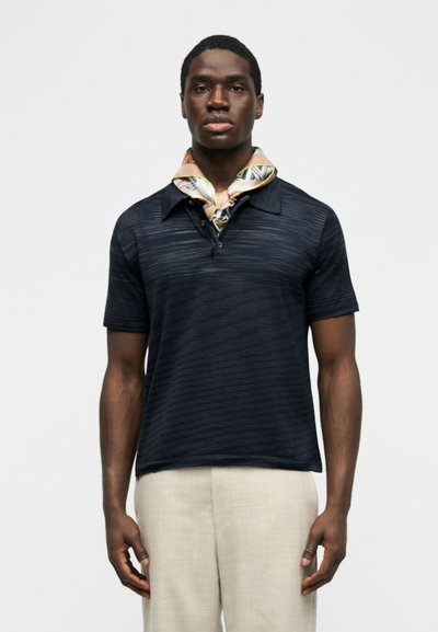 Νέος άνδρας που φορεί σκουρόχρωμο πόλο με κοντά μανίκια, μπεζ παντελόνι και μεταξωτό φουλάρι με σχέδιο δεμένο γύρω από τον λαιμό του, μπροστά σε απλό φόντο.