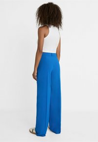 Blaue weit geschnittene Hosen aus glattem Stoff, mit hoher Taille und minimalen Details, kombiniert mit einem weißen ärmellosen Oberteil.