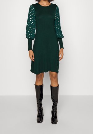 Woman wearing a dark green knee-length dress with gold polka dot puff sleeves and black knee-high patent leather boots.