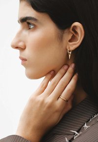 Persona joven con cabello oscuro que lleva pendientes dorados en forma de picos y un anillo dorado, tocándose el cuello, vestida con una chaqueta de rayas.