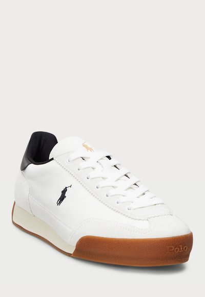 Vit sneaker med ovandel i mocka och läder, svart hälkappa, klassisk snörningsdesign och brun gummisula med präglad logotyp.