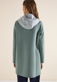 Femme aux longs cheveux portant un sweat à capuche vert à manches longues avec une capuche grise, debout de dos devant un fond beige uni.