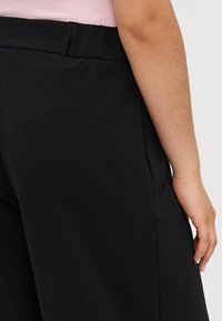 Calças largas pretas feitas de tecido suave, com uma cintura oculta e bolsos laterais, apresentando uma vista de perto das costas e da mão.