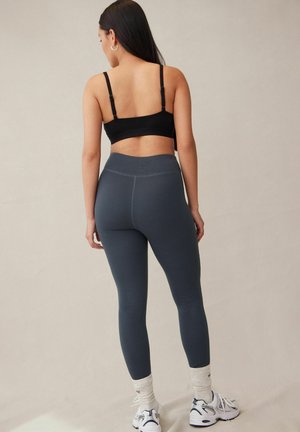Pelēkas leggingas ar teksturētu audumu, augstu jostasvietu, apvienotas ar melno krūšturi un baltām zeķēm, skatītas no aizmugures, stāvot uz neitrāla fona.
