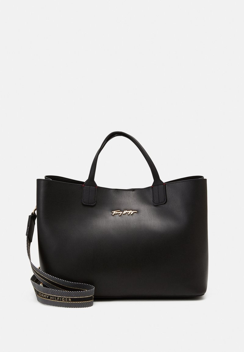 Sac cabas en cuir noir avec deux poignées, une bandoulière rayée amovible avec le texte "Tommy Hilfiger" et un petit logo doré sur le devant.