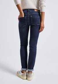 Jean en denim bleu foncé à coupe slim, avec des poches arrière et des poignets retroussés, associés à des baskets blanches. Texture lisse avec un design classique.