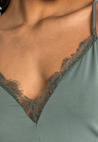 Grön linne med V-ringning, med delikat spetsdetalj längs ringningen och justerbara axelband. Slät och mjuk textur.