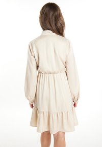 Beige, pitkähihaisessa mekossa on kapeampi vyötärö ja levenevä hame. Suuri tekstuuri, röyhelöhelma ja klassinen kauluksen muotoilu.