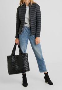 Person iført en sort quilted jakke, lys grå skjorte, blå jeans, sorte hælede støvler, holder en stor sort vævet tote taske.
