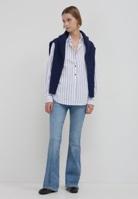 Gilet sans manches tricoté marine sur une chemise blanche à boutons avec des rayures noires, associé à un jean bleu clair évasé et des chaussures noires.