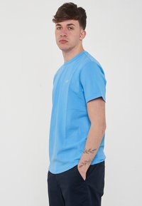 T-shirt di cotone azzurro chiaro con maniche corte, scollatura rotonda e un piccolo logo verde nella parte sinistra del petto. Tessuto morbido, vestibilità casual.