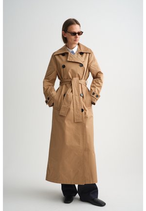 Naine kannab pikkade mustade nuppudega beežikat põlvikut, musti pükse, musti jalatseid ja päikeseprille, seisab ühevärvilise valge tausta ees.